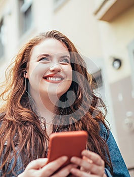 Smiling red curly long hair caucasian teen girl walking on the street and browsing the internet using the modern smartphone.