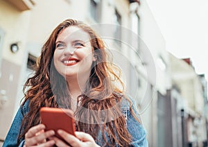 Smiling red curly long hair caucasian teen girl walking on the street and browsing the internet using the modern smartphone.