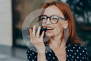 Smiling red-haired business woman using virtual digital voice assistant while standing outdoors