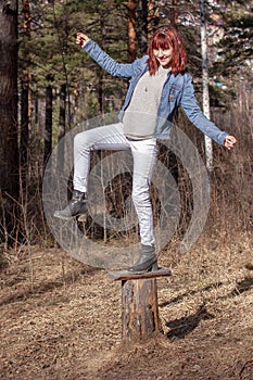 Smiling red-haired teen girl fooling around standing on one leg on a stump in the forest.