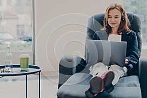 Smiling red-haired woman working in headphones on laptop computer in modern open plan office