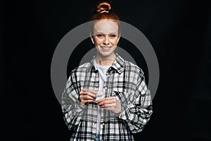 Smiling red-haired young woman in wireless earphones looking at camera on isolated black background