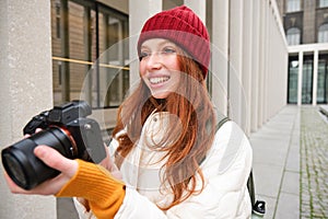 Smiling redhead girl photographer, taking pictures in city, makes photos outdoors on professional camera