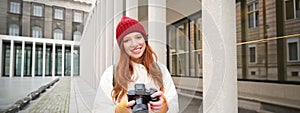 Smiling redhead girl photographer, taking pictures in city, makes photos outdoors on professional camera