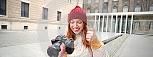 Smiling redhead girl photographer, taking pictures in city, makes photos outdoors on professional camera