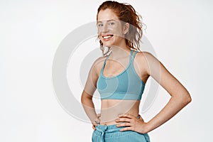 Smiling redhead sportswoman in sportsbra, hold hands on waist and looks happy doing sports. Fitness girl workout against