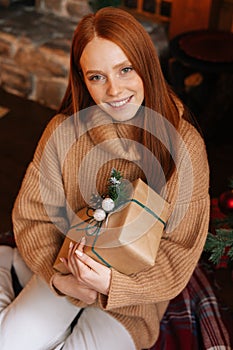 Smiling redhead woman looking at camera happily holding gift box in hand received for Christmas.