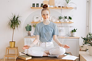 Smiling redhead young woman is meditating while sitting on desk at home office, looking at camera.