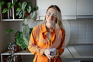 Smiling relaxed woman plant lover in orange dress holding mug in cozy kitchen, look at camera