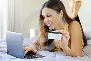 Smiling relaxed young woman lying on bed insert credit card number on notebook  doing safely shopping online at home