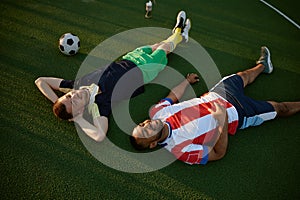 Smiling restful two football players lying on green field grass overhead view