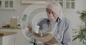 Smiling retired man reading book drinking tea enjoying leisure time in kitchen at home