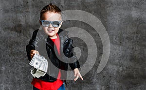 Smiling rich kid boy millionaire in sunglasses, leather jacket and red t-shirt handing us a bundle stack of dollars cash