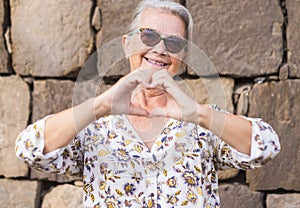 Smiling and romantic senior woman white hair making heart shape with hands expressing love standing against a stone wall with