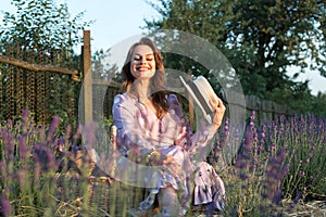 Smiling romantic woman sitting among lavender lit by sunset sunlight. She is wearing a pink dress with a hat