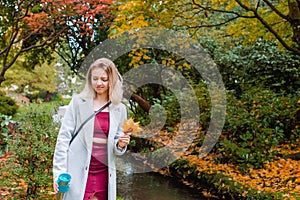 Smiling romantic young woman in coat enjoying a hot drink in her reusable thermos cup, picking fallen leaves during a