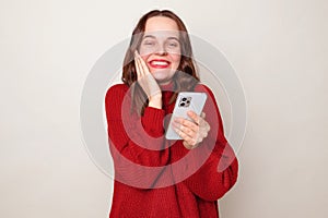 Smiling satisfied Caucasian woman wearing warm red sweater using smartphone isolated over gray background looking at camera with