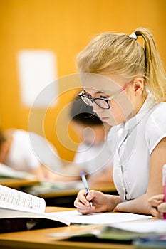 Smiling scholar girl sitting with other children in classroom and writing on textbook. Happy student doing homework at elementary