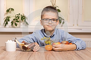 A smiling school-age boy sits at a table and brings fruits to him. With the other hand, he moves the sweets away from him.