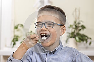 A smiling school-age boy sits at a table and puts a teaspoon of white sugar in his mouth.