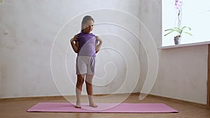 Smiling school age girl in sportswear standing and kneading shoulders with arms bent at elbows on gym mat in empty room