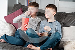 Smiling school aged boys sit on sofa, eat popcorn and watch comic TV show. Kids having fun and relaxing in home room.