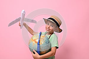 Smiling school boy looking at camera while playing with paper airplane and a globe, standing isolated over pink background with