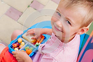 Smiling schoolboy eating his lunch from lunch box sitting outdoor