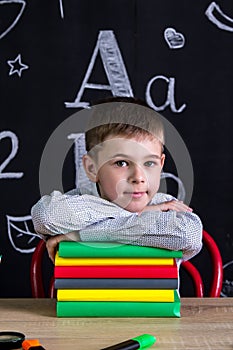 Smiling schoolboy sitting at the desk with a pile of books under the chin, surrounded with school supplies. Chalkboard