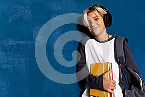 Smiling schoolboy in wireless headphones with textbooks and backpack