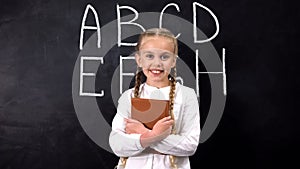 Smiling schoolgirl hugging book, alphabet written on blackboard behind, system