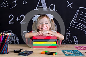 Smiling schoolgirl sitting at the desk with a pile of books under the chin, surrounded with school supplies. Chalkboard