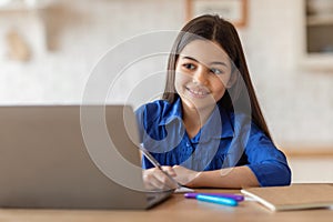 Smiling Schoolgirl Writing Notes Looking At Laptop Studying At Home