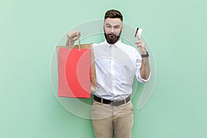 Smiling self-confident businessman showing to camera red shopping bag and credit card.