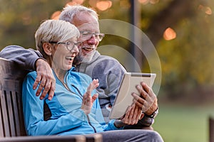 Smiling senior active couple sitting on the bench looking at tablet computer