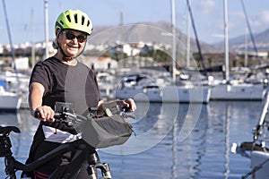 Smiling senior adult woman with yellow helmet standing watching the harbor near her bicycle. Sailboats in the background, blue sea