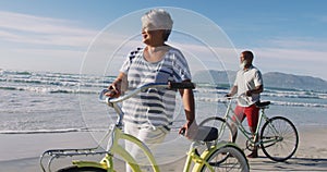 Smiling senior african american couple walking with bicycles at the beach