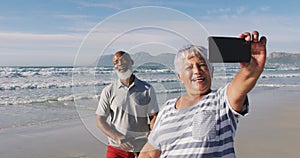 Smiling senior african american couple walking with bicycles and taking a selfie at the beach