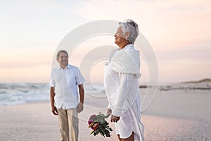 Smiling senior biracial groom looking at bride holding bouquet standing on shore during sunset