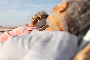 Smiling senior biracial man sitting on chair while looking at woman relaxing on beach
