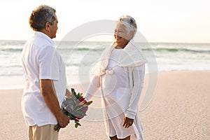 Smiling senior biracial woman holding hand of man with bouquet at beach during wedding ceremony