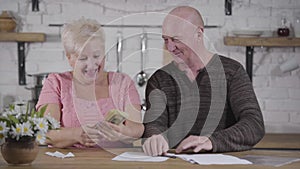 Smiling senior Caucasian man showing pack of banknotes to his shocked wife. Mature woman throwing out bills, taking