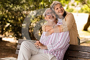 Smiling senior caucasian wife hugging husband, sit on bench, have fun in park outdoor