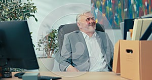 Smiling senior company executive sits in comfortable swivel chair nextcomputer looking around him reminiscing about