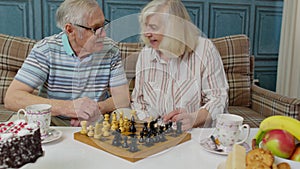 Smiling senior couple grandfather grandmother resting on sofa drinking coffee, playing chess at home