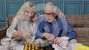 Smiling senior couple grandfather grandmother resting on sofa drinking coffee, playing chess at home