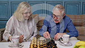 Smiling senior couple grandfather grandmother resting on sofa drinking coffee, playing chess at home