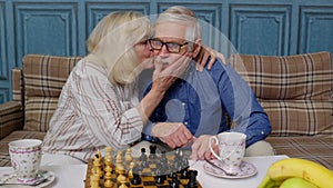 Smiling senior couple grandfather grandmother resting on sofa drinking coffee, playing chess at home