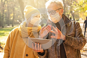 Smiling senior couple in protective mask using digital tablet in autumn park