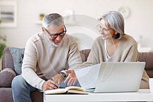 Smiling senior couple reading notification letter with good news from bank while sitting with laptop